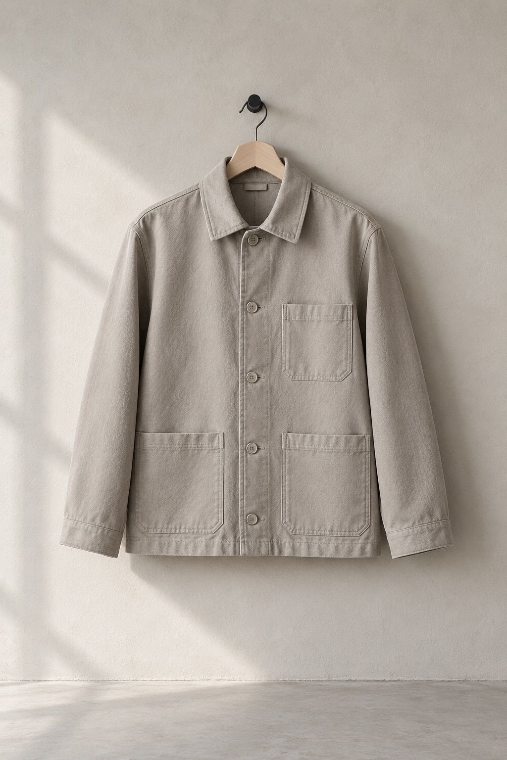 Keld chore jacket in stone on a plain wooden hanger against a pale plaster wall